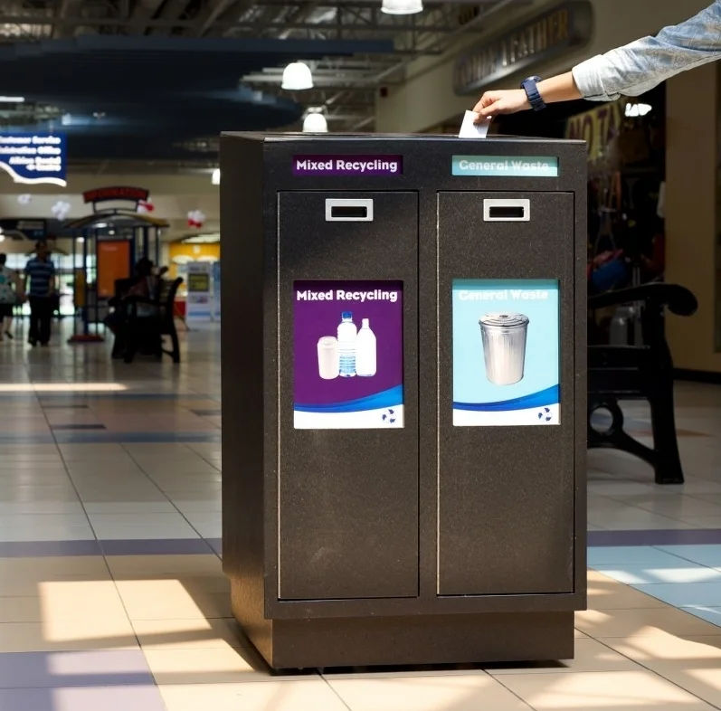 Mall recycling station with two compartments labeled for mixed recycling and general waste, with a person using it